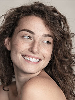 Close up portrait of female model with curly brown hair, brown eyes, freckles and a smiling. Model displays anti wrinkle skin