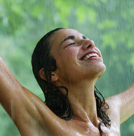 Female featuring dark brown hair standing under the rain outdoors smiling with eyes closed and arms in air